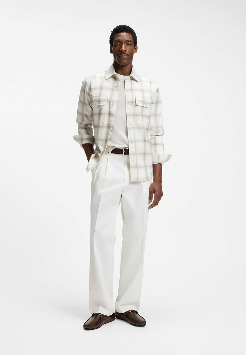 Homme debout, les mains dans les poches, portant une chemise blanche à carreaux sur un t-shirt blanc, un pantalon blanc et des chaussures marron sur un fond blanc uni.