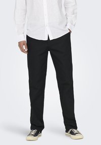 Pantaloni neri in cotone dritti, abbinati a una camicia bianca a maniche lunghe. Le scarpe sono sneakers nere con suole e lacci bianchi.