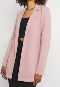 Młoda kobieta ubrana w prążkowany jasnoróżowy żakiet na czarnym teksturowanym topie odsłaniającym brzuch i spódnicy, ozdobiona srebrnym naszyjnikiem z łańcuszka.