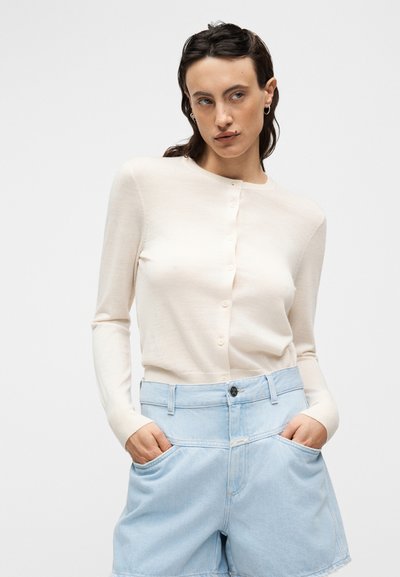 Ανοιχτό μπεζ ζακέτα από απαλό ύφασμα, με στρογγυλή λαιμόκοψη και κουμπιά, συνοδευόμενο από ανοιχτό μπλε τζιν σορτς με μεγάλες τσέπες.