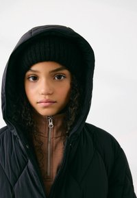 Mustast tikandiga täispuhutav jope suure kapuutsiga. Allpool on pruun tõmblukuga särk. Kootud must müts. Lokkis juuksed raamivad nägu.