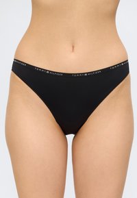 Culotte de bikini noire taille basse avec logo Tommy Hilfiger sur la ceinture, portée par une personne sur un fond uni.