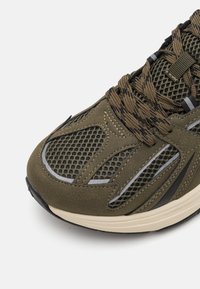 Olive green athletic shoe with mesh and suede materials, featuring a low profile, textured laces, and gray accents on the sides and sole.