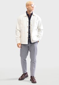 Lichtbeige jas met grote zakken, knoopsluiting en ribgebreide manchetten, gecombineerd met een donker shirt en grijze broek. Bruine schoenen maken de look compleet.