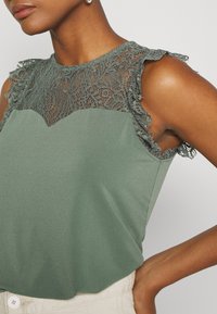 Zielona bluzka z koronkową aplikacją na ramionach oraz falbankami. Gładki materiał, okrągły dekolt i lekko A-liniowy krój.