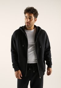 Black zip-up hoodie made from soft fabric, featuring a double hood, front pockets, and drawstring closure. Worn over a white shirt.
