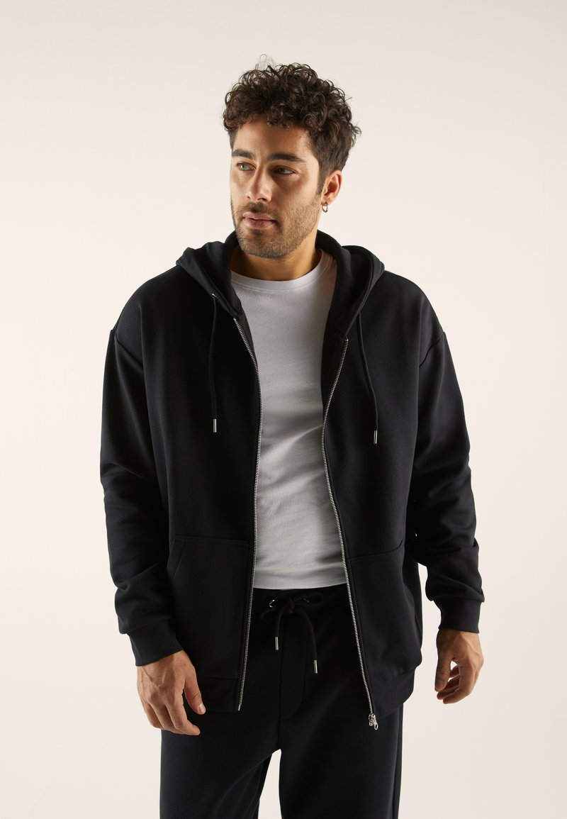 Black zip-up hoodie made from soft fabric, featuring a double hood, front pockets, and drawstring closure. Worn over a white shirt.