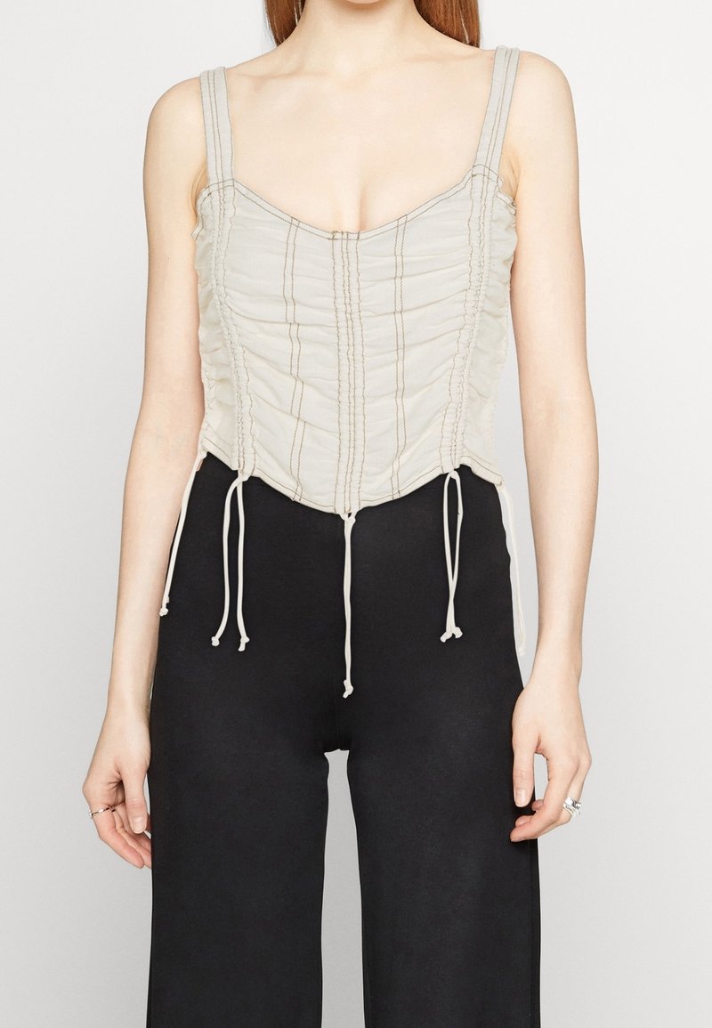 Ljust beige korsettopp med vertikal ruching, justerbara snören i midjan och tunna band. Har sydda detaljer genom hela plagget.