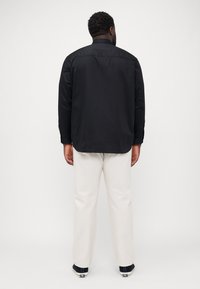 Schwarzes Langarmhemd mit Button-Down-Kragen, glatter Textur und einem Rückeneinsatz, kombiniert mit cremefarbenen Hosen und schwarzen Schuhen.