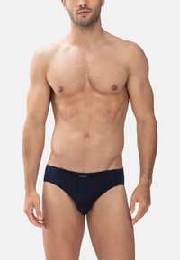 Muskuløs mand iført ensfarvede sorte trusser står mod en hvid baggrund og viser torso og overben, armene afslappet ved siden.