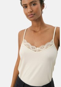 Vaaleanbeige tanktoppi, jossa on ohuet hihnat, pitsireunallinen V-pääntie ja pehmeä, sileä kangas. Mallissa on rento istuvuus.
