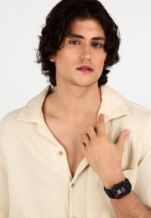 Smartwatch con cinturino nero, quadrante rettangolare che mostra l'ora e le metriche della salute, indossato su un polso nudo; camicia a trama leggera.