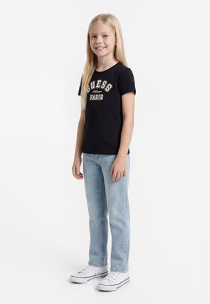 Smilende pige med langt blondt hår iført en sort GUESS Paris t-shirt, lyseblå jeans og hvide sneakers, stående på hvid baggrund.