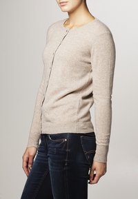 Beige stickad kofta med rund halsringning, knäppning fram och ribbade muddar; bärs över mörkblå jeans, slät textur.