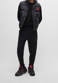 Gilet imbottito nero con un logo rosso, maglietta a maniche lunghe nera, pantaloni neri con coulisse e sneakers nere con dettagli rossi.