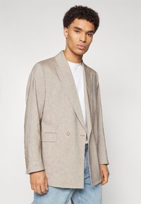 Light beige linen blazer with a relaxed fit, wide lapels, two front buttons, and side pockets, paired with a white t-shirt and faded jeans.