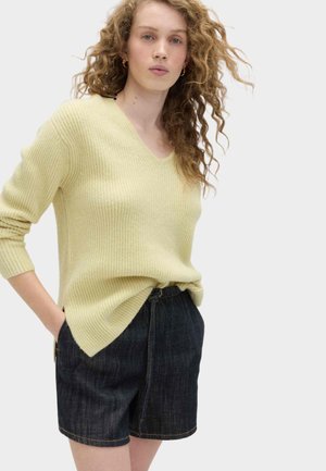 Vrouw met krullend haar, gouden oorringen, een lichtgeel geribd truitje en donkerblauwe denim shorts, met haar handen in de zakken.
