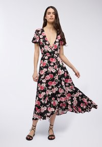 Abito floreale nero con scollo a V profondo, maniche corte a volants e una gonna fluente con un motivo di fiori rosa e bianchi. Indossato con sandali con cinturini.