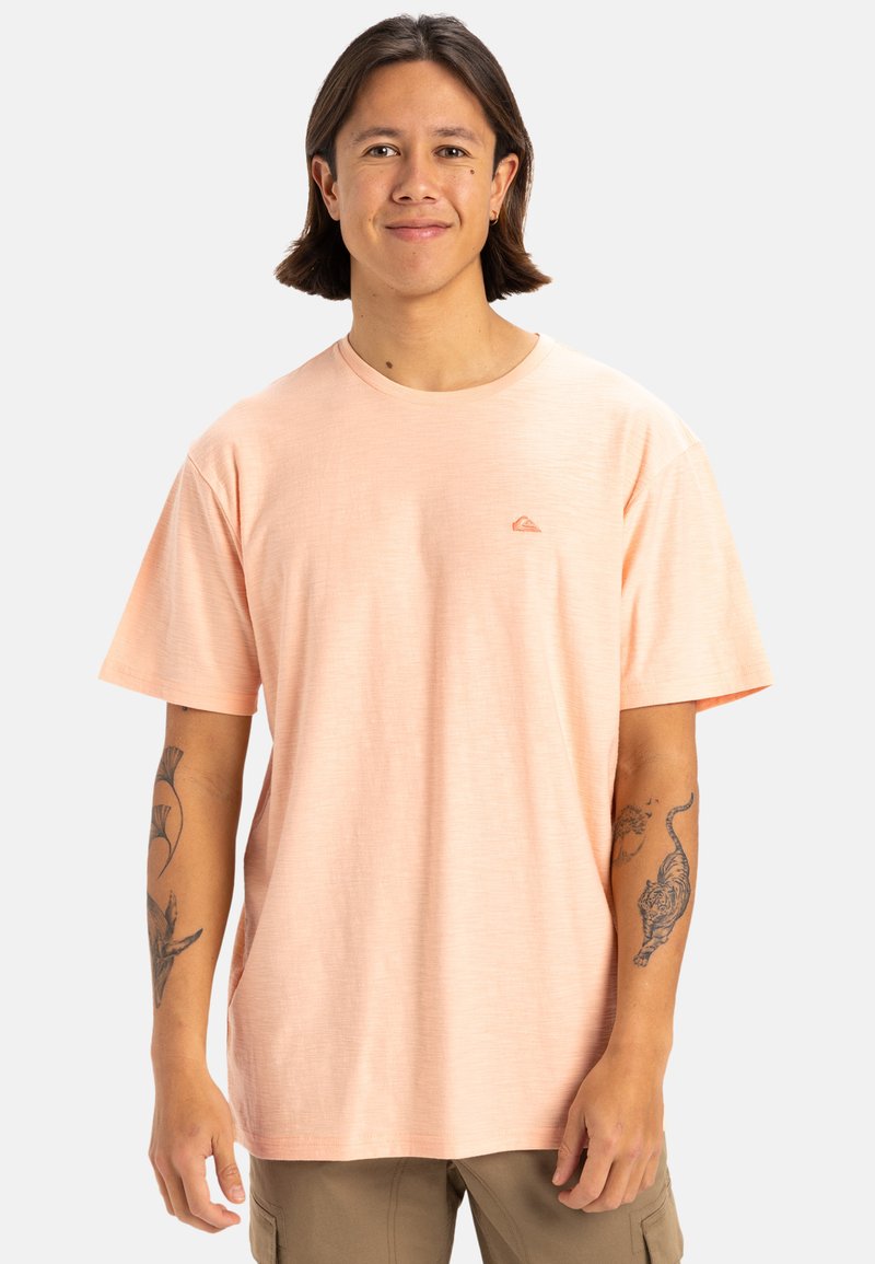 Camiseta de manga corta color melocotón claro con cuello redondo, que presenta un pequeño logo en el pecho izquierdo y una tela suave y texturizada.