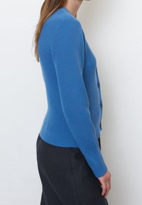 Žena v přiléhavém modrém žebrovaném kardiganu s knoflíky a tmavých kalhotách, stojící z profilu na bílém pozadí.