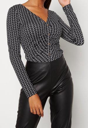 Femme portant un haut à manches longues à motifs noir et blanc avec col en V et boutons, associé à un pantalon en similicuir noir taille haute.