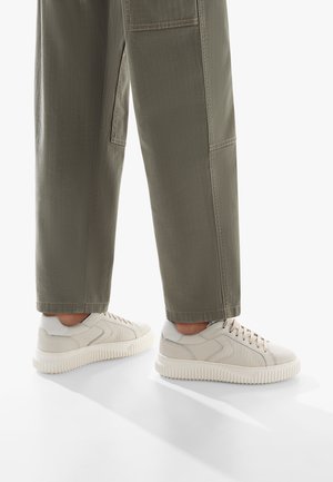 Sneakers beige chiaro in pelle testurizzata con una suola bianca chunky, abbinati a pantaloni verde oliva con cuciture e tasche visibili.