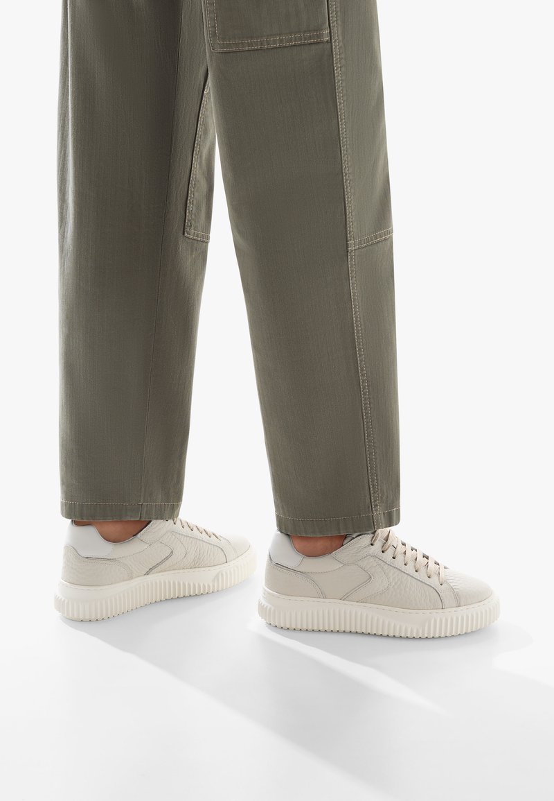 Sneakers beige chiaro in pelle testurizzata con una suola bianca chunky, abbinati a pantaloni verde oliva con cuciture e tasche visibili.