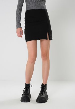 Jambes féminines portant une mini-jupe noire avec une petite fente latérale, un pull gris côtelé et des bottes compensées noires en cuir à lacets.