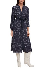 Vestito lungo a maniche lunghe di colore navy con motivi circolari bianchi, scollo a V, vita con cintura, spacco laterale, abbinato a stivali alti bianchi con tacco a stiletto.