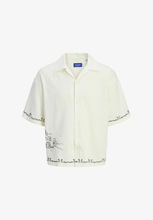 Korte mouwen crème shirt met zwart grafisch ontwerp, gecombineerd met jeansshorts tot de knie. Model draagt witte sokken en zwarte sneakers.