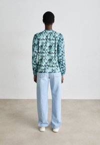 Blus med lång ärm i blågrön mönstring och rynkad halsringning; kombinerad med ljusblå, löst sittande jeans och vita sneakers.
