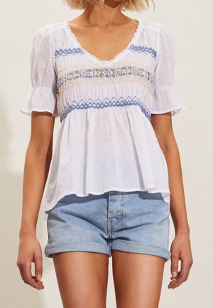 Femme portant un chemisier blanc à manches courtes avec une broderie florale bleue et un short en jean bleu clair roulé.