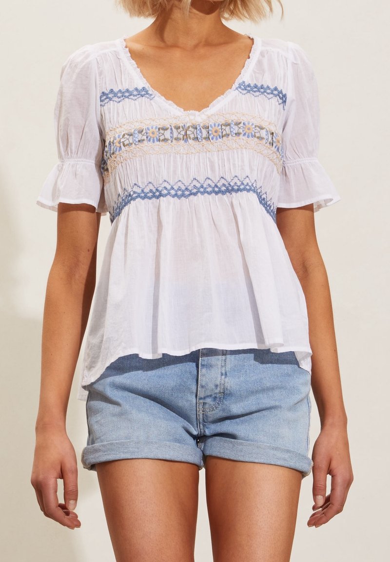 Femme portant un chemisier blanc à manches courtes avec une broderie florale bleue et un short en jean bleu clair roulé.