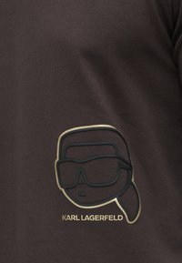 T-shirt castanho escuro com uma imagem de um rosto com óculos de sol em preto e dourado, com o texto "KARL LAGERFELD" abaixo. Textura de tecido suave.