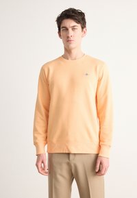 Peach sweatshirt made of soft fabric, featuring a crew neckline and ribbed cuffs. Embroidered logo detail on the chest. Worn with beige trousers.