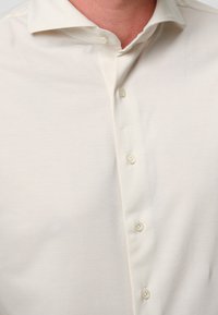 Camicia button-up beige chiaro con colletto classico, caratterizzata da una texture liscia e cinque bottoni. Design semplice, senza motivi o accenti.