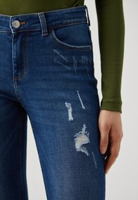 Σκούρο μπλε τζιν παντελόνι με ψηλή μέση, φθορές και σχισίματα. Διακριτές χρυσές ραφές και σχέδιο πέντε τσεπών.