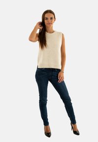 Top in maglia senza maniche di colore crema con orlo a coste, abbinato a jeans skinny blu scuro e tacchi alti neri, su uno sfondo bianco semplice.