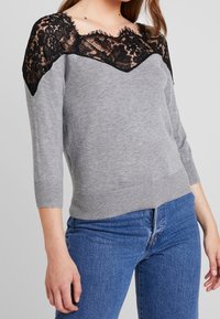 Pull gris avec une superposition en dentelle noire aux épaules, manches trois-quarts et coupe ajustée, associé à un jean en denim bleu.