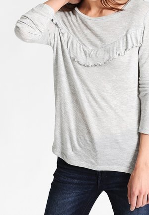 Mujer con una blusa de manga larga gris claro con un detalle de volantes en el pecho, combinada con vaqueros azul oscuro.
