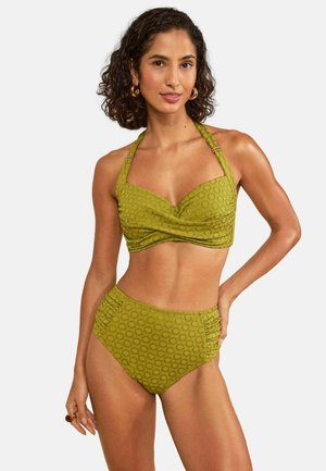 Femme aux cheveux bouclés portant un bikini taille haute vert olive texturé, avec des boucles d'oreilles anneaux dorés et une bague, debout avec une main sur la hanche.