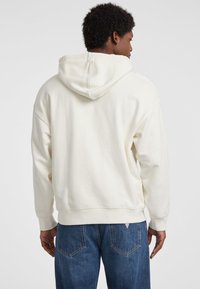 Sudadera de color crema con capucha, de corte relajado, puños acanalados y bolsillo tipo canguro. La tela parece suave y texturizada.