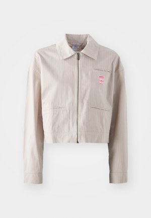 Zkrácená béžová bunda vyrobená z hladkého materiálu, s límcem, předním zipem a malou červenou nášivkou na hrudi. Viditelné vertikální pruhy.