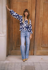 Sinise ja valge mustriga pluus volüümikate varrukatega, kombineeritud heledate laia säärega teksade ning beežide kontsakingadega, taustal puituks.