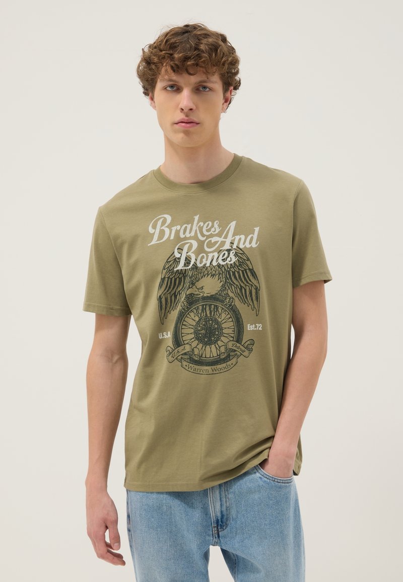 Junger Mann mit lockigem Haar, der ein olivgrünes T-Shirt mit Adler- und Rad-Grafik und dem Schriftzug „Brakes And Bones“ trägt, kombiniert mit hellblauen Jeans.