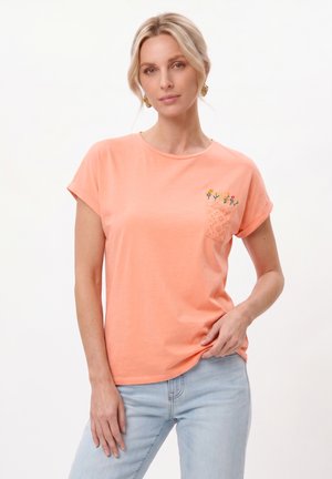 Frau mit blondem Haar trägt ein pfirsichfarbenes T-Shirt mit einem kleinen bestickten Blumenmuster auf der Brusttasche und hellblaue Jeans.