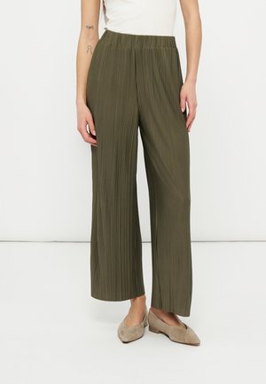 Donna che indossa pantaloni verdi oliva a pieghe e a gamba larga, top smanicato bianco e scarpe basse in suede beige, in piedi su sfondo bianco.