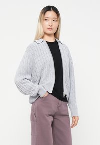 Cardigan grigio a maglia con motivo a treccia, design con colletto e chiusura a zip, indossato sopra una maglietta nera e abbinato a pantaloni color malva.