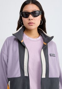 Fleece jacket in lavender and gray with cream accents; features a high collar, full zipper, and logo patches. Layered with a light purple shirt.