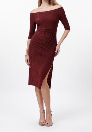 Vrouw draagt een bordeauxrode off-shoulder midi-jurk met geplooide taille en hoge split, gecombineerd met nude enkellaarsjes met bandje.